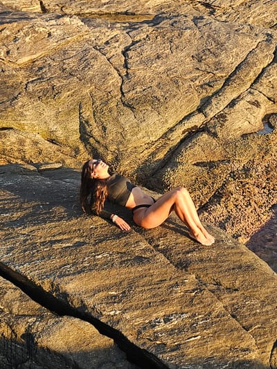 A woman in a black swimsuit lies on tan rocks. Her head is oriented towards the bottom of the frame, with her feet pointed toward the top. She faces the camera. Her body is curved, with her knees bent and her right arm lying next to her hip. There are light brown rocks to the left of her head, with some green moss on the rock above her. The rock to her right is a light tan color with a dark crack in the bottom right corner. The rocks are located in Armação dos Búzios, Brazil.