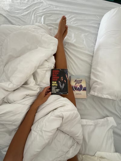 In Khan Mean Chey, Cambodia, a person's legs are visible, draped over white bedding. Two books lie on the covers: one titled "Женщины, которые любят слишком сильно" (Women Who Love Too Much) by Robin Norwood, and another, "Магия утра" (The Morning Magic). The scene is indoors, likely a bedroom, with a large white pillow to the right of the frame.
