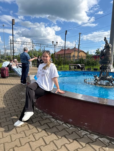 The image, correctly oriented, depicts a bright, sunny outdoor scene in Malaya Vishera, Russia, likely near a train station. A young woman with blonde hair, wearing a white t-shirt, black trousers, and white sneakers, is the central subject. She smiles faintly at the camera, leaning on the dark red edge of a large, ornate fountain. Sunglasses rest on her head.
The fountain features vibrant blue water and intricate dark metal sculptures depicting human-like figures and possibly marine life, with water jets creating splashes. In the background, several other people are visible; one man in a blue patterned shirt stands with his back to the camera, gesturing, while others are seated on benches, some next to a red suitcase.
The ground is paved with interlocking grey concrete pavers. Above, a clear blue sky with scattered white, fluffy clouds indicates bright, mid-day sunlight. Numerous railway overhead power lines and support structures, along with streetlights, span the scene, confirming the proximity to a railway line. A building with an orange-red roof and a decorative black fence are also visible in the distance. No legible text is discernable.