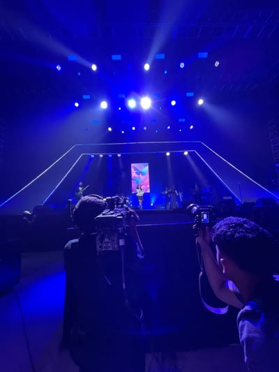 The image depicts a concert stage in Salembaran, Indonesia. The stage is illuminated with blue lights and features a large screen displaying a performer. Several cameramen are positioned to capture the event, focusing their lenses on the stage. The environment is dark except for the stage lighting, highlighting the performers and the technology used to film them.