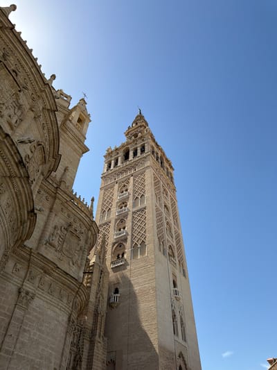 This media file is a low-angle shot of the Giralda bell tower and the Seville Cathedral in Seville, Spain. The sky above is a clear, bright blue, indicating a sunny day, likely in the late afternoon or early evening given the warm, golden light illuminating the stone structures and casting gentle shadows.
The subjects are the impressive architectural features of the cathedral and its iconic bell tower. The Giralda, a former minaret, is a towering, ornate structure made of brick, adorned with intricate geometric patterns and windows. It is seen from below, emphasizing its height and grandeur. In the foreground, the detailed stonework of the cathedral's facade is visible, featuring elaborate carvings, sculptures, and decorative arches. Small gargoyle-like figures can be seen perched on the roofline. The overall scene conveys a sense of historical significance and architectural splendor.
There are no people visible in the frame, and no specific activity or event is taking place; the focus is solely on the static, monumental architecture. The details of the stonework, the clear sky, and the warm lighting are the most notable elements. The location is clearly indicated as Seville, Spain, known for this architectural masterpiece. The mood is one of awe and appreciation for the historical and artistic value of the structures. There is no visible text in the image.