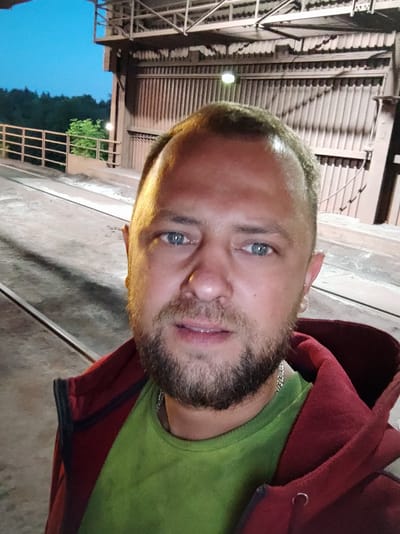 A man with light brown hair, blue eyes, and a beard takes a selfie outdoors at what appears to be an industrial site in Salzgitter, Germany. He is wearing a green t-shirt and a red hooded jacket. The background shows industrial structures with corrugated metal siding and a clear blue sky. Railway tracks are visible on the ground in front of the man.