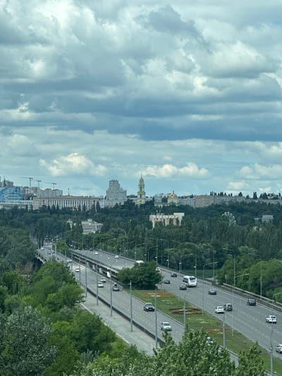 This is a high-angle, full-shot photograph of a multi-lane highway cutting through a densely wooded area, with a cityscape in the background. The highway, which appears to be part of the Vologodskaya Highway in Lipetsk, Russia, curves from the lower left towards the upper right of the frame. Numerous cars and a bus are visible, traveling in both directions on the highway. The road is lined with streetlights and surrounded by lush green trees.

In the background, a prominent cityscape unfolds. A large, stately building with white columns and a flag flying on top stands out. To its left, a modern glass building with a blue facade is under construction, with several cranes visible. To the right of the stately building, a tall, slender bell tower with a yellow dome pierces the sky, and further to the right, a golden-domed church can be seen. A Ferris wheel is also partially visible amidst the trees in the middle ground, suggesting a park or recreational area. The sky is filled with dramatic, gray clouds, indicating overcast weather.