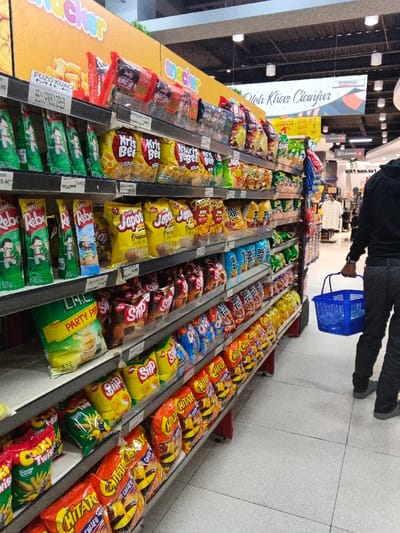 A well-stocked grocery aisle displays various snack items, including potato chips from brands like "Kris Bee," "Japota," "Chitato," and "Siip." Other snacks like "Rebo" are also present. The aisle is organized with products neatly arranged on shelves. A shopper with a blue shopping basket is seen in the aisle. Signage above the chips section reads "Oleh Khas Cianjur" and "Snacker," while a smaller sign indicates prices and promotional offers.