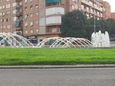 The image depicts a street scene in Badajoz, Spain, featuring a fountain and a building. In the foreground, a section of a road is visible with a curb and a lush green grassy area. Beyond the grass, a multi-tiered fountain sprays water upwards and outwards, creating arcs of water. The fountain appears to be located in a circular or semi-circular formation.
Behind the fountain, a building with a brick facade rises several stories. The building has numerous windows, some with balconies and awnings. A business with a sign reading "orcopplata" is visible at the ground level of the building. Trees are present behind the fountain, with their foliage forming a green backdrop. A street lamp with blue accents is also visible, positioned in front of the building and behind the fountain. The sky above is overcast, suggesting a cloudy day. Several parked cars can be seen in the background, hinting at a residential or commercial area. No people are directly visible in the image, though the presence of cars and businesses suggests an urban environment.
