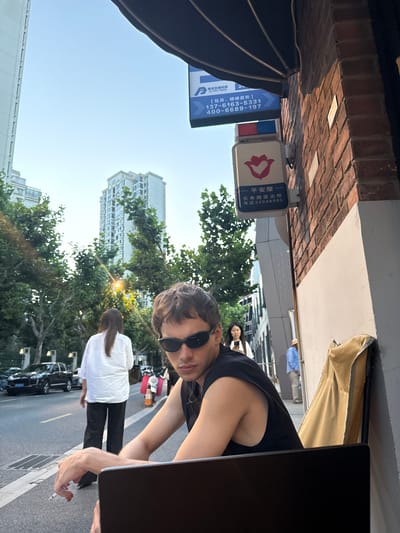 This image captures an outdoor urban scene during the golden hour, indicating late afternoon or early morning, with a clear blue sky and bright sunlight. The setting appears to be a street or public area in Putuo District, China, surrounded by tall city buildings and green trees.

In the foreground, a person with short dark hair and sunglasses, wearing a black sleeveless top, leans into the frame from the right. They are holding a lit cigarette. A black laptop is partially visible next to them. To the far left, a large black photography umbrella or softbox is present, suggesting a photoshoot or filming activity. Further back on the street, another person with long dark hair, dressed in a white shirt and black pants, stands facing away from the camera. Other blurred figures are visible in the background, along with parked cars.

Overhead, a brick wall and a white awning-like structure are visible. Several signs are prominent, featuring Chinese text. One blue sign displays "杨杰交通科技" (Yang Jie Transportation Technology) and "CJ TFC TECH," along with phone numbers "137-6163-5331" and "400-6689-197" for "Monthly, Off-peak Consultation." Another sign with a red hand logo reads "平安屋" (Safety House / Peace House), indicating "长寿路派出所" (Changshou Road Police Station) with the phone number "电话:22048085."

The overall atmosphere suggests a casual, possibly candid moment during an outdoor activity, likely related to photography or videography, taking place in a busy urban environment.