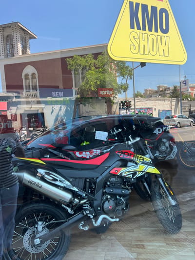 The image captures a reflection on a storefront window, showing a street scene in Bari, Italy. Dominating the foreground are two motorcycles, one a black and red Aprilia with yellow accents, and another partially visible behind it. The Aprilia motorcycle features a prominent silver exhaust pipe with the "aprilia" logo. Through the window's reflection, a building with a distinctly decorated facade and arched windows is visible. A large yellow triangular sign with the words "KMO SHOW" in blue lettering hangs in the upper portion of the frame. Also visible are reflections of traffic lights, buildings with graffiti, a silver car, and a parked rental car. The ground appears to be paved with wooden-style tiles.