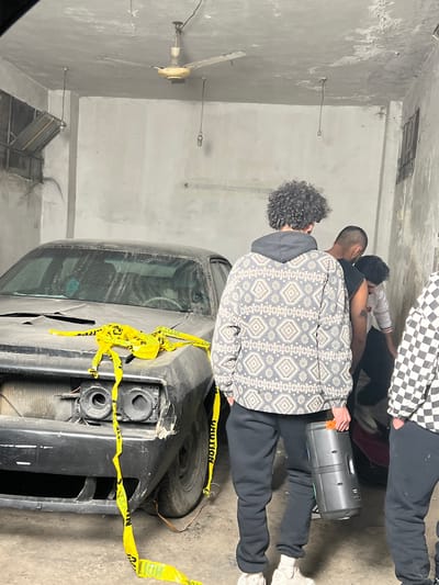 An indoor scene shows an old, damaged black car wrapped in yellow caution tape. Several people stand nearby, including one with curly hair wearing a patterned jacket and another with a checkered shirt. A gray boombox rests near their legs. The room is stark, with a gray wall, a ceiling fan, and exposed electrical fixtures. The atmosphere suggests an informal gathering amidst a vehicle investigation or accident scene.