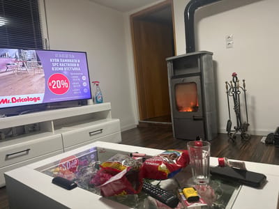 The image captures an indoor living space in Krauchenwies, Germany. In the foreground, a white coffee table with a glass top holds a black Samsung remote control, a black smartphone, an empty clear glass, a pink disposable lighter, a yellow disposable lighter, an opened pack of Marlboro cigarettes, and multiple open snack bags containing items such as sunflower seeds and pretzels.
To the left, a large flat-screen television rests on a white entertainment unit with drawers and open shelves. The television displays an advertisement for "Mr. Bricolage" with Cyrillic text, offering a discount on laminate and SPC flooring. A blue spray bottle is positioned on the entertainment unit to the right of the TV. A window with horizontal blinds is visible on the far left.
To the right of the scene, a gray, freestanding wood-burning stove with a black flue pipe extending upwards is active, with visible flames behind its glass door. Adjacent to the stove, a decorative black metal stand holds a set of fireplace tools. A double electrical outlet is present on the white wall above and to the right of the stove. A dark wooden door frame leading into another room is visible behind the stove. The room features dark wooden flooring and plain white walls. No individuals are present in the scene.