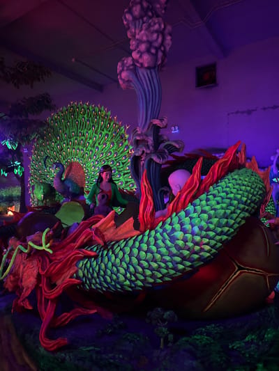A vivid, neon-lit scene in Binh Lu, Vietnam, presents a surreal tableau. A green-scaled dragon with red tendrils surrounds a bald baby figure. Adjacent to this is a peacock sculpture with vibrant green and blue feathers. Underneath the peacock are assorted plants and structures. The entire scene is illuminated with purple and blue lighting. In the background, a picture frame and some other objects are attached to the wall.