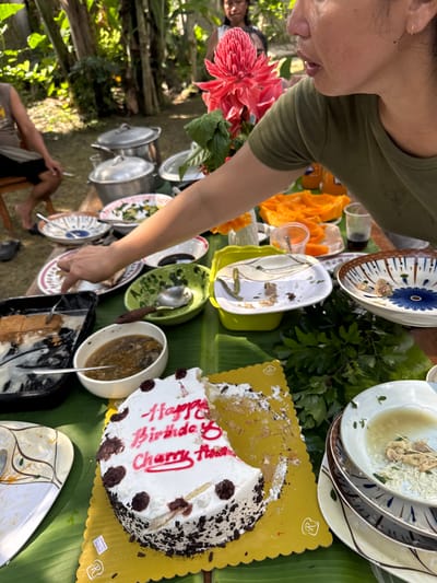 An outdoor celebratory meal is depicted in Carmen, Philippines, taking place on a surface covered by large green banana leaves. In the foreground, the upper right portion of a person wearing a green t-shirt is visible, with their right arm extended towards the table, suggesting serving or reaching for food. To the far left, a person is seated on a wooden stool, partially visible. In the background, lush green foliage and trees indicate a tropical environment, with bright sunlight casting dappled shadows.

The table features an array of dishes and serving items. Prominently in the lower foreground is a partially eaten round white cake with chocolate sprinkles, resting on a yellow scalloped cake board. Red icing on the cake reads "Happy Birthday Charry Tho." To the left of the cake is a dark rectangular tray containing what appears to be squares of food with a white topping, some of which have been consumed. Adjacent to this is a white bowl holding a brown, stew-like dish with visible vegetables or mushrooms.

Further into the frame, numerous plates and bowls are scattered, some containing food remnants, others appearing nearly empty. These include decorative ceramic plates, a light green plastic bowl with residual green leafy matter, and a small bowl with a dark liquid. A stack of used, patterned plates is visible on the far right. In the background, several large silver pots with lids are present, indicating a substantial meal. Slices of orange fruit (possibly papaya) are arranged on a white tray, and plastic bottles of an orange beverage are also visible, along with clear plastic cups. A vibrant red torch ginger flower (Etlingera elatior) with green leaves is positioned centrally, partially obscuring a person's face in the middle ground.