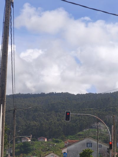 The image depicts a street scene in Moaña, Spain. In the foreground, a wooden utility pole stands on the left, with electrical wires running across the top of the frame and horizontally across the middle ground. Further into the scene, a two-way traffic light is visible, with one light displaying red. Below the traffic light is a building and further behind, a forested mountain covers the background. Above the mountain, a cloudy sky is visible. There is a pedestrian crossing sign near the bottom. Several buildings with red tiled roofs are seen scattered on the mountainside.
