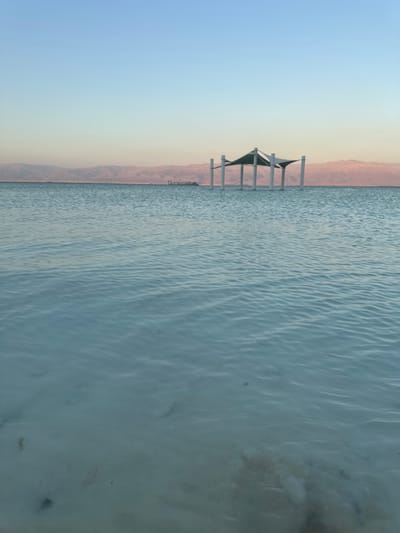 This is a vertical, eye-level, medium shot of the Dead Sea at dusk in Tamar Regional Council, Israel. In the foreground, the shallow, calm, light blue water shows a sandy bottom with scattered rocks and debris. The middle ground features a modern, square-shaped canopy structure supported by four white pillars, rising out of the water. In the background, distant, hazy, pink-hued mountains form a stark horizon line against a soft gradient sky that transitions from a pale pink near the mountains to a light blue at the top of the frame. There are no people or animals visible in the image.