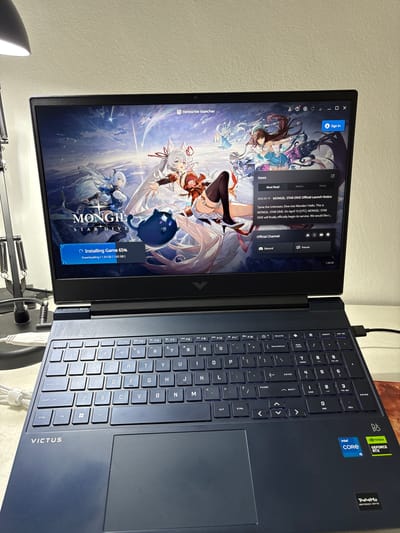 A dark blue HP Victus laptop is open, oriented with the screen facing towards the left side of the frame. The screen displays an anime-style illustration with the title "MONGIL STARDIVE" and an installation progress bar showing 65% completion. An open window on the screen shows game-related news and channel options. The keyboard is visible on the right side of the laptop, along with Intel Core i5 and NVIDIA GeForce RTX stickers. A black cable is plugged into the top right side of the laptop. The laptop is situated on a white surface, with a partially visible lamp arm and base in the lower part of the frame. The background is a light cream or off-white wall. There is no indication of Córdoba, Argentina.