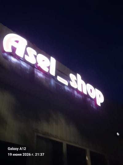 An outdoor, low-angle, night shot captures a bright, illuminated sign displaying "Asel_shop" in white, stylized lettering against a dark, slightly blurred background. The letters appear to have a subtle pink or purple glow around their edges. Below the sign, at the bottom left of the image, a timestamp and device model are visible: "Galaxy A12" and "19 июня 2026 г. 21:37". The overall environment suggests an urban setting at night, possibly a storefront or commercial establishment in Kara-Köl City, Kyrgyzstan. No people are visible in the frame.