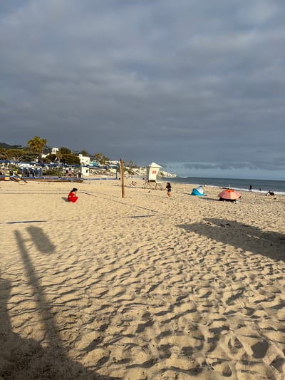 A wide shot captures a sandy beach in Laguna Beach, California. The foreground is dominated by rippled sand casting long shadows, indicative of late afternoon or early morning sun. In the mid-ground, a volleyball net stretches across the frame, with a lone figure in a red outfit crouching near its center. Several seagulls are scattered across the sand. Further back, a lifeguard tower stands prominently. A couple of beach umbrellas, one orange and white striped and another blue, are set up on the sand. Various people are visible enjoying the beach in the distance. The ocean forms the background, with gentle waves lapping the shore. The sky is overcast with dark, dramatic clouds, suggesting an approaching storm or the end of a day. Buildings and palm trees line the coastline in the distance, typical of the Laguna Beach landscape.