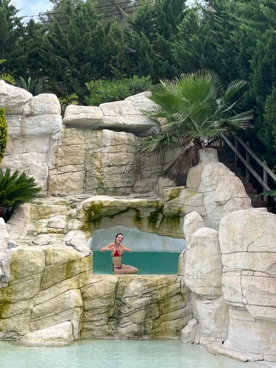 In Can Güell, Spain, a woman in a red bikini is seated in a pool of clear, turquoise water. She is positioned within a man-made rock formation that simulates a waterfall, with water visible behind her. She is smiling and giving a double thumbs-up gesture. The rock formation is made of light-colored, textured material, appearing to be artificial stone. Lush green trees and foliage form the backdrop, and a wooden staircase is visible to the right, leading upwards alongside the rock structure. The pool itself has light-colored tiling or lining at the bottom.