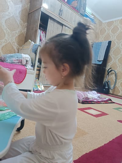 A young girl with her hair in a bun, wearing a white outfit, is seated at a blue table, her hands near a collection of green plastic packets. She is positioned in a room with patterned wallpaper and a large wooden wardrobe. A vacuum cleaner is visible in the background, near a doorway. A rug with geometric patterns covers the floor. The scene appears to be an indoor residential setting in Navoiy City, Uzbekistan.