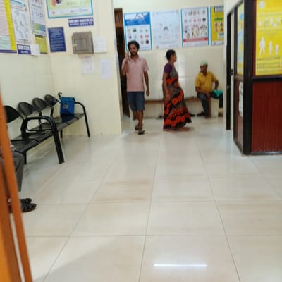 The image depicts the interior of a waiting room in Marna, India, possibly a clinic or hospital. The room has a tiled floor and painted walls, with informational posters displayed on the walls. There is a row of black metal chairs with a blue bag placed on one. Three people are visible: a man is walking while talking on a mobile phone, a woman in a patterned sari is walking, and an elderly man in a yellow shirt is sitting on a bench inside a doorway. The room is well-lit and appears to be clean.
