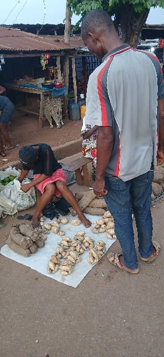 The image captures a street scene in Abakaliki, Nigeria, at coordinates 6.343, 8.106. A woman, wearing a black shirt and pink skirt, sits on the ground. She is engaged in selling produce, specifically yams and other root vegetables, which are spread out on a white sheet in front of her. A man, wearing a grey t-shirt with red stripes and blue jeans, stands to the right of the woman. His arms are crossed, and he appears to be observing the woman's business. Behind the woman and to the right, a partially obscured individual, likely another vendor, sits on a wooden chair, possibly also selling produce. The environment includes a dirt road, a concrete sidewalk, and a building with a rusty corrugated roof. Various items and structures are present in the background, including a small wooden stand, a variety of bags and baskets, and a tree. The scene is well-lit and suggests a bustling marketplace atmosphere.