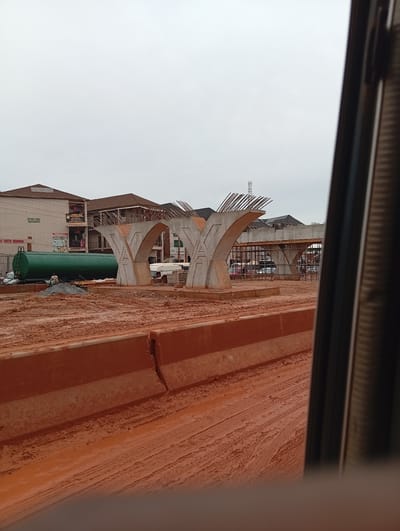 This image depicts an ongoing construction site in Katambe, Nigeria. The scene is dominated by a partially constructed elevated road or bridge, characterized by large concrete pillars with exposed rebar. In the foreground, red soil covers the ground, with tire tracks indicating vehicular movement. Orange concrete barriers line the visible roadway. In the mid-ground, a large green cylindrical tank is visible next to a fenced building. The building appears to be a commercial structure with signage. Several vehicles, including a white van and other cars, are present in the background near the construction. Two individuals are near the green tank, one appearing to be working. The sky is overcast.
