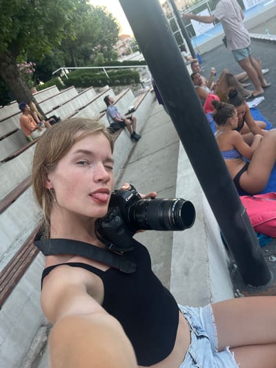 In Gošići, Montenegro, a young woman with blonde hair and freckles is taking a selfie in an outdoor amphitheater. She is wearing a black tank top and denim shorts, and she is holding a Canon camera. She is sticking her tongue out and winking at the camera. In the background, several other people are sitting on the amphitheater's tiered seating, some of whom appear to be sunbathing. The environment is lush with trees and greenery, and there is a bright, sunny sky overhead.