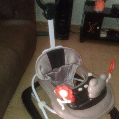 The scene depicts an indoor domestic setting in the city of Abakaliki, Nigeria. A light-brown and black baby walker, featuring a padded seat and a play tray, occupies the foreground. The play tray includes a red and white floral toy, a light-colored animal-shaped toy, and a red and white circular hanging toy attached via a white stem. The walker has white support rods and a black base, with a black handle extending upwards from the rear. In the blurred background to the left, a section of a brown upholstered sofa is visible. To the right, a dark-colored side table with a gold "LV" logo supports an orange decorative plant. A tall black speaker stands beside the table. The floor is light-colored and tiled, with visible cables near the speaker and table. No individuals are present in the image.
