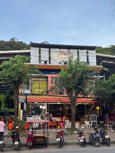 The image depicts a street scene in Ban Klong Haeng, Thailand. The foreground is dominated by a row of shops and restaurants lining the street. Several motorcycles are parked along the sidewalk, indicating active commerce. One person in a pink shirt is visible standing on the sidewalk, facing away from the camera. The buildings are a mix of styles, with some featuring colorful awnings and decorative elements. Trees are interspersed along the street, providing some shade.

The shops appear to be open, with goods displayed outside and visible through windows. The overall impression is that of a bustling commercial area. The time of day appears to be daytime, likely late afternoon, given the shadows and lighting. The weather seems clear with a bright sky visible in the upper left portion of the image.

Notable details include a circular mural on one of the buildings, featuring an intricate design with figures and patterns. Various items are displayed outside the shops, including furniture, decorations, and what appear to be food or drink items. Visible text on a license plate of a motorcycle reads "101 5840". Another license plate reads "101 502". A sign above a shop entrance has some Thai script. The overall atmosphere is lively and suggests a typical day in this Thai locale.