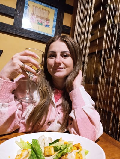 A woman is seated at a wooden table in what appears to be a restaurant. She holds a wine glass in her left hand and is touching her hair with her right. She is wearing a pink and white jacket. In front of her is a white plate of salad. Behind her are decorative golden strands hanging vertically. On the wall in the background is a framed picture of three pigs. The location is Chernogolovka, Russia.