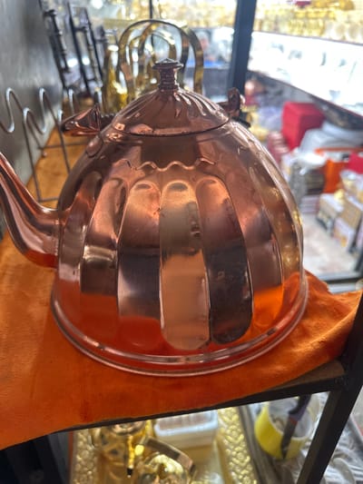 The image shows a close-up of a polished copper teapot with vertical ribbed detailing. The teapot is resting on an orange cloth, which is on a shelf. In the background, there are blurred displays of goods, suggesting a market or shop setting. Specifically, the shelves in the background appear to contain various items, possibly decorative or functional, with some golden accents visible. The lighting is bright, reflecting off the shiny surface of the teapot. The environment appears to be indoors, possibly a bazaar or a retail store in Kara-Suu, Kyrgyzstan, known for its large market where a wide variety of goods are sold.