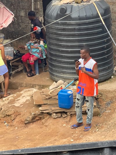 An outdoor scene shows a man in an orange jersey looking at his phone next to a large water tank. A blue container with a hose rests beside him. In the lower left corner, people are seated on a low structure. Another person reclines on the ground. The surroundings appear modest, with a dirt ground and weathered materials.