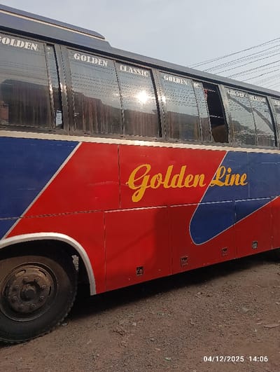 The image displays the side of a large passenger bus, located in Kaliganj, Bangladesh. The bus features a red lower body panel with "Golden Line" written in yellow script, and an upper blue section with "GOLDEN CLASSIC" in white text repeated across its windows. The windows are tinted, and one window panel appears partially open. Sunlight reflects prominently off one of the tinted windows. The front left wheel, consisting of a black tire and rim, is visible in the lower left portion of the image. The bus is situated on an unpaved surface composed of dirt and small debris. Overhead power lines are visible against the light sky in the background. No individuals are present in the scene. A digital timestamp "04/12/2025 14:06" is visible in the lower right corner.