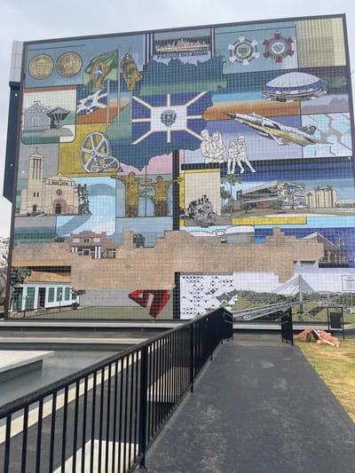 The image depicts a large, multi-paneled tile mural covering the facade of a building at Praça do Expedicionário (Expeditionary Square) in the city center of Anápolis, Goiás, Brazil. The mural illustrates various historical, cultural, and economic aspects of Anápolis and the state of Goiás.

The mural features numerous distinct mosaic panels. Visible elements include:
*   **Upper section:** Portraits of historical figures, the Brazilian and Anápolis flags, the Anápolis municipal crest on a map of Goiás, the "Parque das Águas" text, and the logos for "SEMTUR" and "SEBRAE."
*   **Mid-section:** Depictions of a church, a large industrial gear, a monument with two figures, a family group, a fighter jet, a train, and modern buildings including a stadium and what appears to be an airport control tower.
*   **Lower-mid section:** A significant portion of the mural in this area appears to be incomplete or under renovation, showing a lighter brown, textured surface where tiles are absent or discolored, obscuring parts of underlying architectural depictions.
*   **Lower section:** Portions show a traditional green-roofed building, a stylized red geometric "1" shape, white circular patterns, and a cable-stayed bridge.

In the foreground, a black metal railing extends along the left, bordering a dark paved pathway that leads towards the mural. To the left of the pathway is a rectangular water feature or pool with light-colored concrete edges. To the right of the pathway, a manicured green lawn is visible, on which a light brown statue of a dog, lying down, is situated. The sky above is overcast. No people are actively depicted in the foreground or engaged in specific actions.