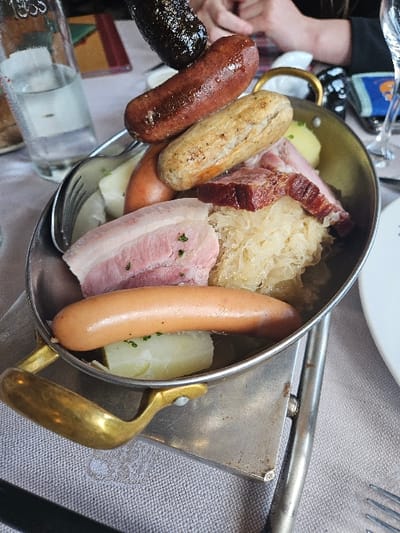 A metal pan with a brass handle sits on a metal stand, atop a white linen tablecloth. It contains a traditional Alsatian dish known as "Choucroute Garnie," consisting of a variety of meats, potatoes, and sauerkraut.  The meats include a large slice of pork belly, two thick slices of smoked ham, a long sausage, a few smaller sausages, and a black pudding. The potatoes are quartered, and the sauerkraut is coarsely chopped. The pan is resting on a metallic stand with a small metal plate on top, next to a fork and a white ceramic plate. The scene appears to be set in a restaurant or cafe, as evidenced by the partially filled glass of water and a wine glass in the background, indicating a dining establishment. The location is in the town of Scherwiller, France, based on the provided coordinates.