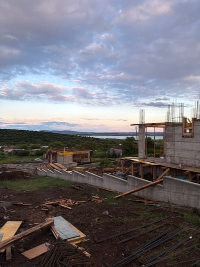 The image shows a construction site in Cherni Vrah, Bulgaria, against a backdrop of a sunset-lit sky. The partially constructed building features concrete walls with exposed rebar and wooden framework for windows and doors. The surrounding landscape includes a forested area leading to a body of water in the distance. Construction materials like lumber and rebar are scattered on the ground around the building's base. There are no people visible in the scene.