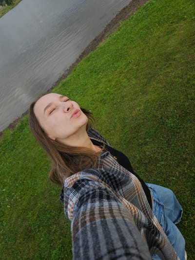 A young woman with brown hair is taking a selfie outdoors. She is wearing a plaid shirt and jeans. Her eyes are closed, and her lips are puckered as if she is about to kiss. She is standing on a grassy area next to a body of water. The environment suggests a park or recreational area, possibly near the Kama River in Elovo, Russia, given the proximity to water and open green space. The lighting is overcast, indicating a cloudy day.