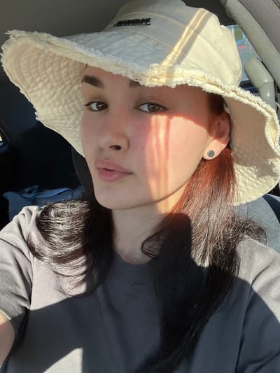 A young woman with dark hair and brown eyes wears a light-colored bucket hat with frayed edges. She is inside a vehicle, likely in the city of Starobaltachevo, Russia, with sunlight casting diagonal shadows across her face and clothing. She is looking towards the camera, and her expression is neutral. Her attire consists of a dark gray t-shirt. A small, circular earring is visible on her right ear. The background suggests the interior of a car, with a dark seat and what appears to be a gear shift lever.