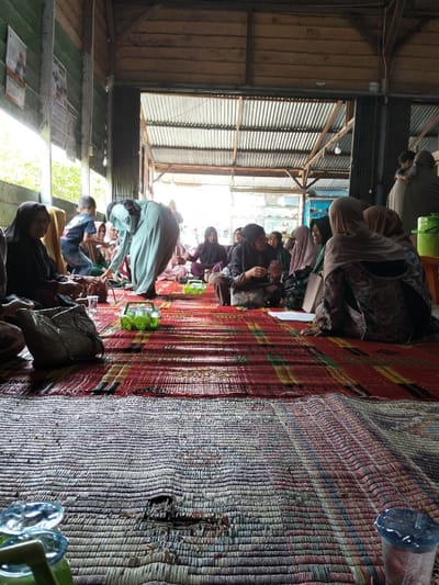 A group of women, mostly wearing hijabs, sit on a mat in what appears to be a community gathering. They are positioned on colorful mats of different weaves. Some women are engaged in conversation, while others are focused on activities in their hands. A child wanders among them. The location seems to be a covered outdoor or semi-outdoor space with a corrugated metal roof and wooden walls. Sunlight streams in from an opening at the far end of the space. Cups and containers are placed on the mat near the seated women.