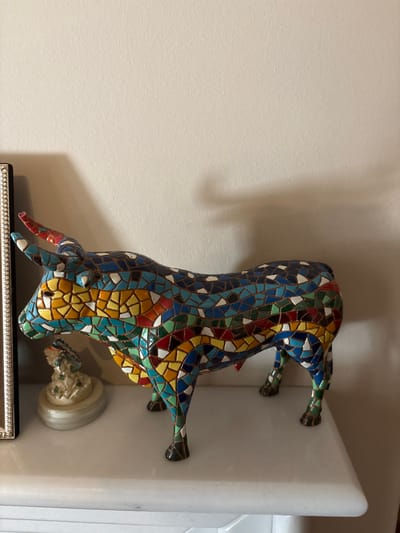 A mosaic bull figurine is displayed on a white shelf against a light-colored wall. The bull is intricately decorated with small, colorful tiles in shades of blue, yellow, red, green, and white, forming a vibrant mosaic pattern across its body. It is positioned with its hind legs resting on the shelf and its front legs extended as if walking or leaping. The bull's head is lowered, and its horns are visible. To the right of the bull, a small, white figurine, possibly a cherub or statue fragment, is also present on the shelf. The shelf itself appears to be made of a white, solid material, possibly marble, with a rounded edge. The background is a plain, textured wall, with a shadow cast on the right side, suggesting indoor lighting. The scene is static, with no apparent activity taking place beyond the display of the objects. The overall impression is one of decorative art within an interior setting. No visible text is present in the image.