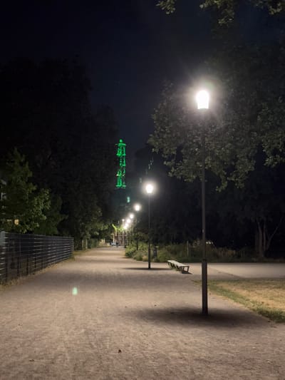 The image shows a path in a park at night. The path is illuminated by streetlights. There are trees on both sides of the path. In the distance, a tall structure is visible, lit with green lights. The scene is in Duisburg, Germany, at the Landschaftspark Nord. The park is a former industrial site that has been transformed into a recreational area. The green-lit structure is likely a part of the former industrial equipment, such as a blast furnace or a cooling tower, that has been artistically illuminated. The atmosphere is quiet and serene.