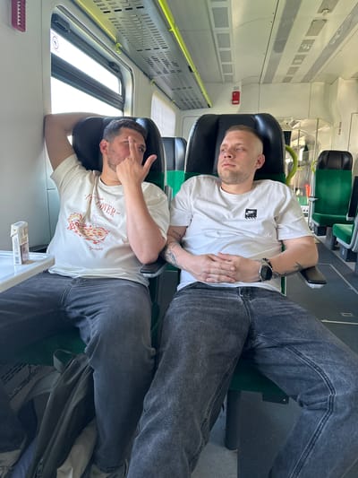Two men are sitting in train seats in Верцялішкаўскі сельскі Савет, Belarus. The man in the foreground is wearing a white t-shirt with a pizza graphic and grey jeans. His right hand is resting on the headrest, and his left hand is raised, covering his left eye. The man in the background is wearing a white t-shirt with a logo and grey jeans, with a watch visible on his left wrist. The train carriage is mostly white and grey with green seat backs.