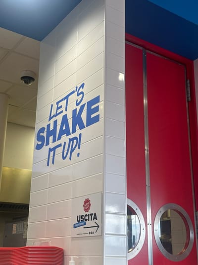 The image depicts an interior scene within a commercial establishment, specifically a Johnny Rockets restaurant located in Arese, Italy. A prominent white tiled column occupies the left and central foreground, bearing large blue text that reads, "LET'S SHAKE IT UP!" Below this text, a white rectangular sign displays a red circular "Johnny Rockets" logo, the word "USCITA" (Italian for "EXIT") in black, a blue line stating "TORNA A TROVARCI" (Italian for "COME VISIT US AGAIN"), and a black arrow pointing to the right. Smaller text below reads, "Condividi la tua JR Experience" followed by "000".
To the right of the tiled column are two bright red doors, each featuring a silver vertical handle/trim and two circular porthole-style windows with silver frames. These doors appear to be commercial swing doors, likely leading to a kitchen or staff area, indicated by the adjacent "USCITA" sign. The ceiling above these doors is painted a solid blue. In the upper left background, a white dome-shaped surveillance camera is mounted on a lighter-colored ceiling. In the lower left corner, a stack of red rectangular trays or plates is visible, along with the top of a white pump dispenser. No individuals are present in the scene.
