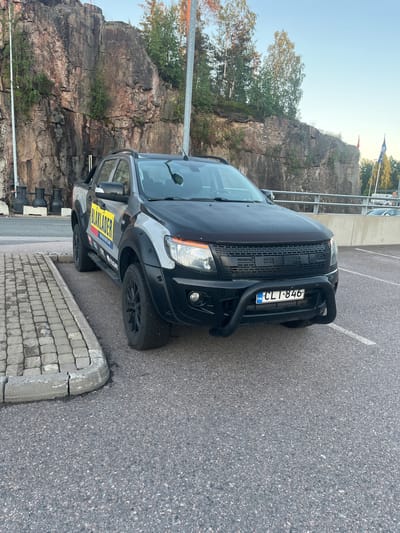The image shows a black Ford Ranger pickup truck parked in a parking lot with a rocky cliff face in the background. The truck has a large yellow decal on the side that says "BLAKLADER WORKWEAR". The license plate of the truck is "CL1-846" and it has a bull bar on the front. The parking lot is paved with asphalt and there is a concrete curb with some paving stones next to it. The background shows a rock formation with some trees growing on it and a clear blue sky. The scene appears to be in Vantaa, Finland, as indicated by the license plate.