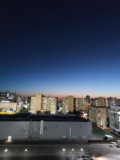 The image depicts a twilight scene of a city with multiple buildings and a gradient sky. The sky transitions from a deep, dark blue at the bottom to a lighter blue and then a faint pinkish hue at the top. A single, bright star is visible in the dark blue portion of the sky.
The cityscape is dominated by several multi-story buildings, appearing to be apartment complexes or office blocks. Many of the buildings have illuminated windows, suggesting that it is evening and people are inside. Some of the buildings are lighter in color, like beige or cream, while others are darker gray. One prominent building on the right side of the image is a large, gray structure with visible pipes and ventilation units on its exterior. Adjacent to this building is a dark paved area, likely a parking lot or road, with several lampposts casting light.
A white car is parked in the paved area. Further down the paved area, what appears to be a pedestrian crossing is marked with white lines. Another vehicle, partially visible, is driving on the road, its headlights illuminating the area.
The scene suggests a time of day just after sunset, given the twilight sky and illuminated buildings. The location is identified as Aktau, Kazakhstan. There are no people clearly visible in the image. The overall mood is calm and urban. Some text is visible on a building in the lower part of the image, appearing to be in Cyrillic script, with letters like "ВНА" and "АЛИА" discernible.