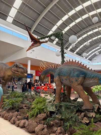 This media file captures an expansive indoor scene, likely a shopping mall or large public concourse, adorned with elaborate dinosaur exhibits in San Francisco Ocotlán (Ocotlán), Mexico. The image is oriented vertically (or 90 degrees clockwise from its natural horizontal perspective).

In the foreground, a large, highly detailed statue resembling a Tyrannosaurus Rex stands on the right side, its head tilted upwards, mouth slightly agape to reveal its teeth. Beside it, to the left and slightly behind, is a massive, quadrupedal dinosaur statue. This dinosaur features a blue-green scaly body with prominent brown-orange plates and spikes running down its back. Its robust legs are textured, blending into a landscaped base of artificial rocks, ferns, and other lush green foliage designed to mimic a prehistoric environment.

Suspended from the high, arched ceiling, a pterodactyl-like flying reptile statue hangs, seemingly in mid-flight, attached by a vine-wrapped structure that also features artificial leaves. The ceiling itself is a striking architectural feature, composed of a lattice of beams with numerous skylights allowing natural daylight to flood the space. Several large, white, spherical light fixtures are also suspended from the ceiling at varying heights.

In the mid-ground and background, a bustling public area is visible, suggesting a food court or common seating zone. There are numerous tables with orange and green chairs, occupied by several people who are sitting, standing, and walking. They appear to be casually observing the dinosaur displays or engaged in conversation. The atmosphere seems relaxed and bright, indicating daytime. Visible architectural elements include white walls, orange pillars, and large windows that offer glimpses of a clear blue sky outside.

Visible text is limited but includes partially obscured letters like "AltruH" on an orange pillar near the T-Rex and what appears to be "S H O P" further in the background. A blue screen on a pillar displays an abstract image. The scene conveys a sense of wonder and entertainment, characteristic of a family-friendly attraction within a modern commercial setting.