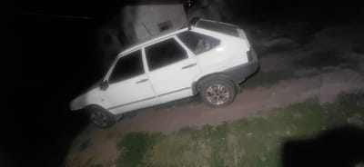 A white hatchback vehicle is parked on a dirt road at night, with a building visible in the background. The car is angled, with its front pointing downwards and slightly to the left. The scene appears to be in the city of Ketrysanivka, Ukraine. The lighting is dim, suggesting it is dark outside. The ground is uneven, with patches of grass and dirt.