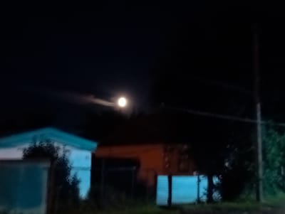 The image depicts a nighttime scene in Qobda, Kazakhstan. The primary subject is the moon, a bright, luminous orb in the dark sky, partially obscured by wispy clouds. Below the moon, silhouetted against the night, are the outlines of several structures, possibly buildings or sheds. One structure in the foreground on the left is painted in shades of white and light blue, with a darker, possibly wooded area obscuring part of its facade. To its right, another structure appears to be a darker, reddish-orange color, with a fence or barrier in front of it. Another section of a building, also in a blue hue, is visible towards the right of the image. The scene is dimly lit, with the moon providing the main source of illumination. The overall mood is quiet and somewhat mysterious due to the darkness and the indistinct nature of the structures. There is no visible text in the image.