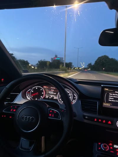 The image shows the interior of an Audi car from the perspective of the driver. The dashboard features a speedometer displaying 102 km/h, a temperature reading of 20.0°C, and a digital display showing song information: "Title: Bad Boys", "Artist: Emanuell", "Album: ", "Play time: 0:14". The car is on a road with streetlights visible in the distance, suggesting it is evening or twilight. Trees line the side of the road. The sky is a deep blue. The car is likely in Marten, Bulgaria, as indicated by the context provided.