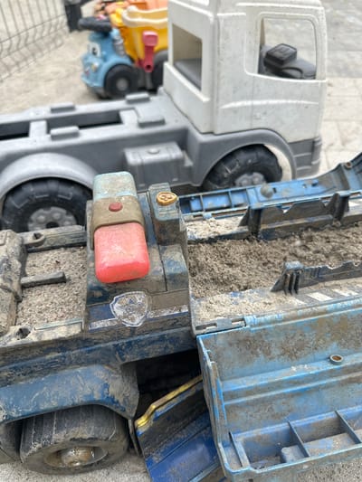 In Malinovka, Russia, a close-up, eye-level shot captures a collection of heavily used and sand-filled toy trucks outdoors. The foreground is dominated by a blue and gray toy truck, its cargo bed filled with sand and debris. A red and blue flashing light assembly is prominently featured on its roof, showing signs of wear. To the left of this truck, a portion of a large gray toy truck is visible. In the background, a small, blue and yellow toy truck with a smiling face is partially visible. The ground appears to be a light-colored, textured surface, possibly concrete or pavement, with a faint grid pattern visible. The overall impression is one of an outdoor play area where these toys have been actively engaged.