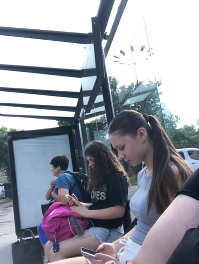 The image shows a group of young people at a bus stop, likely in Chernihiv, Ukraine. A teenage girl with her hair in a ponytail is wearing a grey tank top and white shorts, looking down at her phone. To her left, another young woman with curly hair is wearing a black t-shirt and denim shorts, also engrossed in her phone. In the background, an older woman in a blue shirt and dark pants is seated, looking to her side. She is wearing a backpack. A bright pink backpack with a blue keychain is visible in the foreground. The bus stop has a metal frame and glass panels, with a billboard on the left side. Trees and a white vehicle are visible in the background through the glass. The sky is bright and overcast.